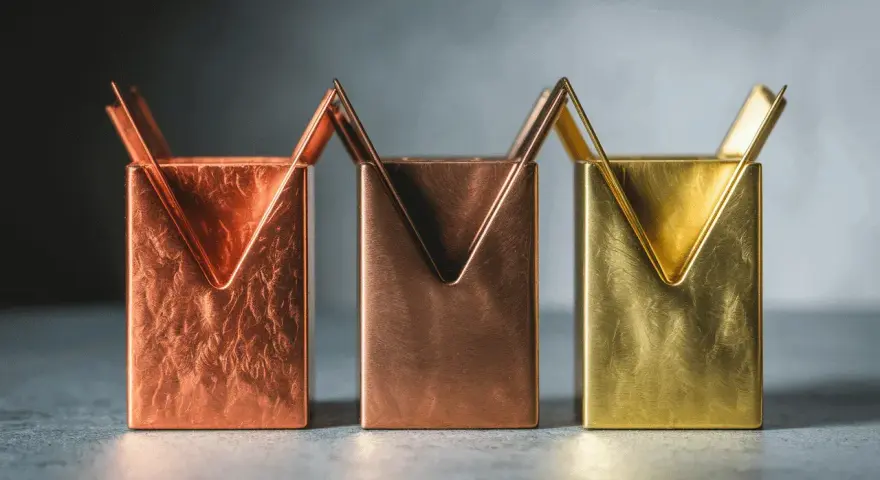 Trois porte-stylos aux formes géométriques, dont un en couleur laiton, sur un fond sombre.