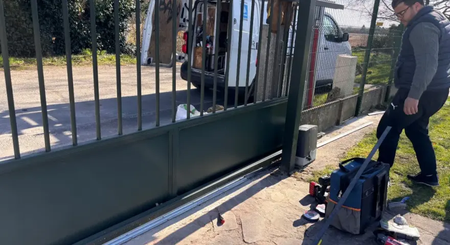 Un homme installe le moteur d'un portail coulissant. Des outils sont posés au sol près de la cellule.