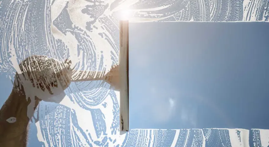 Nettoyage d'une vitre avec une raclette et de la mousse. Le soleil brille à travers la surface savonneuse, créant des reflets. Idéal pour enlever la colle sur vitre.