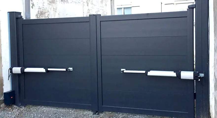 Portail gris à deux battants avec motorisation. Les bras de la motorisation portail sont fixés sur un mur perpendiculaire.