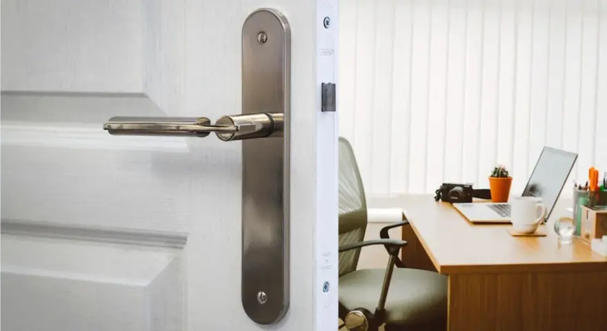 Poignée de porte en métal sur une porte blanche, révélant un bureau avec un ordinateur portable. La serrure BCC, un gage de sécurité.
