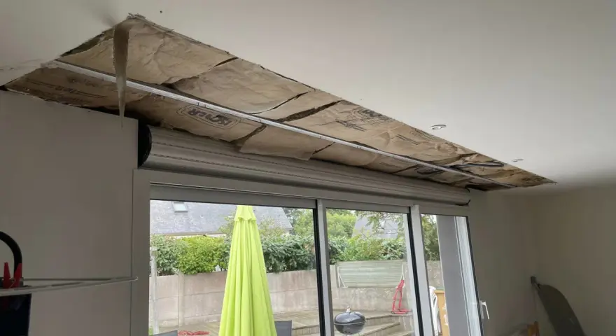 Travaux d'isolation phonique fenêtre PVC coffre : le plafond est ouvert, révélant l'isolant. Dehors, un jardin avec parasol vert.