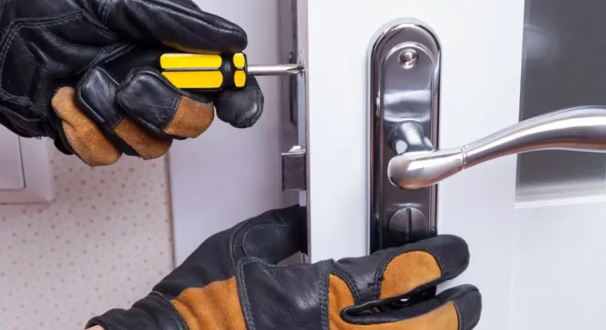 Mains gantées ajustent un loquet serrure bloqué sur une porte blanche avec un tournevis.