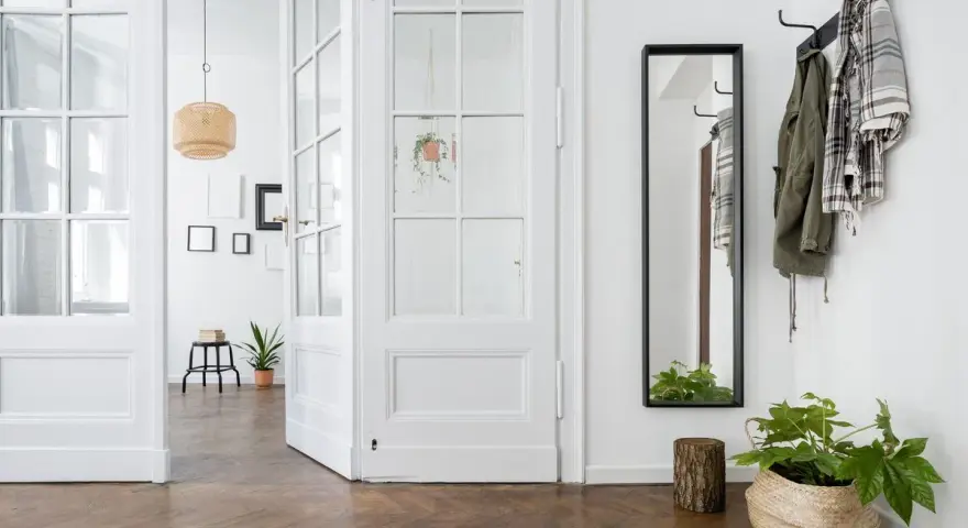 Un couloir lumineux avec une porte vitrée blanche ouverte, un miroir, des manteaux sur un porte-manteau et des plantes. Idéal pour un projet de relooker porte.