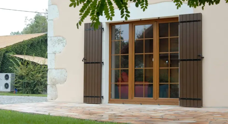Fenêtre en bois avec volets bruns, matériaux menuiserie de qualité. Le jardin verdoyant et la façade en pierre complètent le tableau.