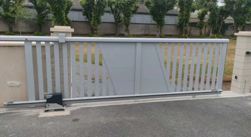 Portail électrique gris coulissant, avec son moteur discret, assure la protection du domicile. Des arbres taillés bordent la propriété.