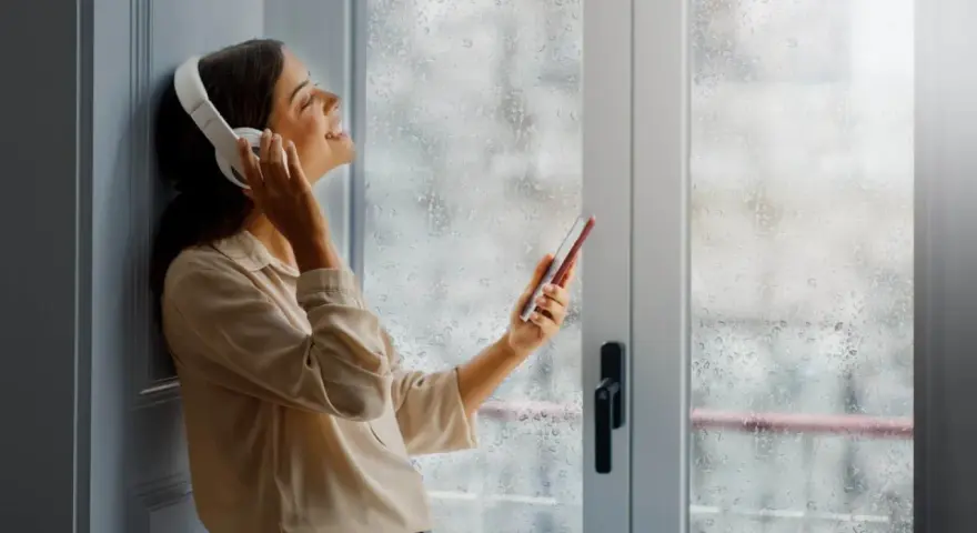 Une femme écoute de la musique avec des écouteurs, profitant du calme malgré la pluie. Un film isolant phonique pour fenêtre pourrait améliorer cette quiétude.