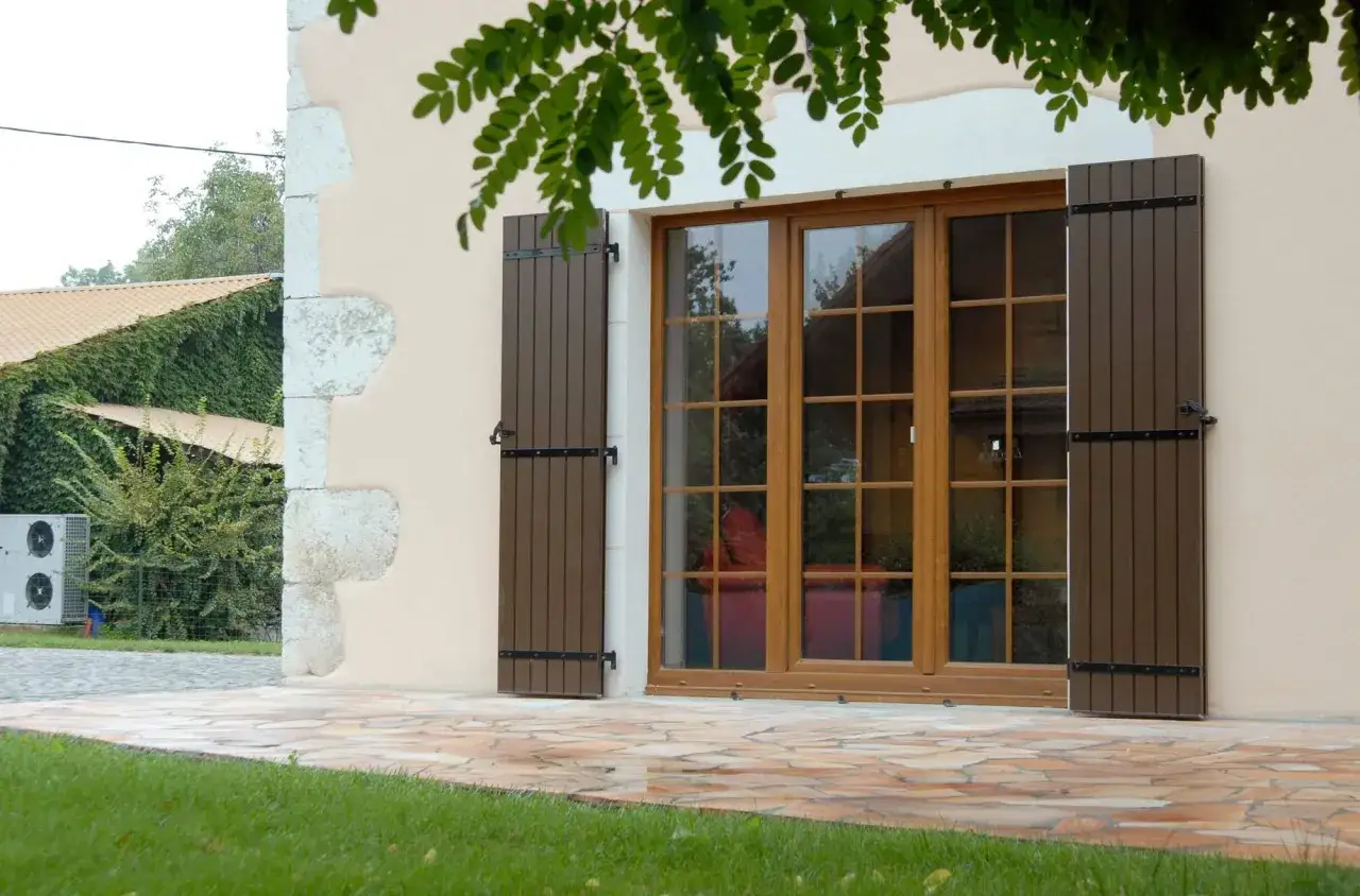 Une belle fen&ecirc;tre en bois ou PVC, avec des volets marron, s'ouvre sur un jardin verdoyant.
