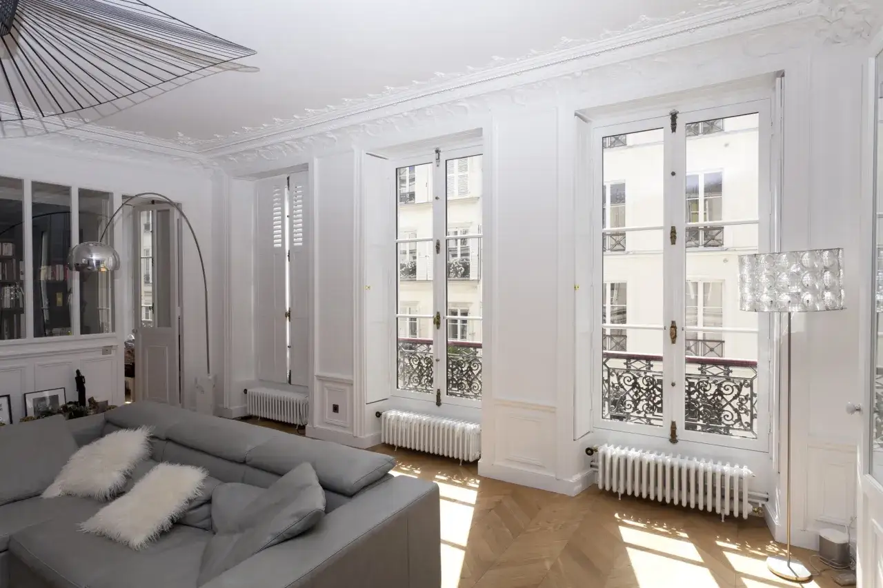 Lumière traversant la fenêtre d'un immeuble parisien, éclairant un parquet en chevrons et un canapé gris.
