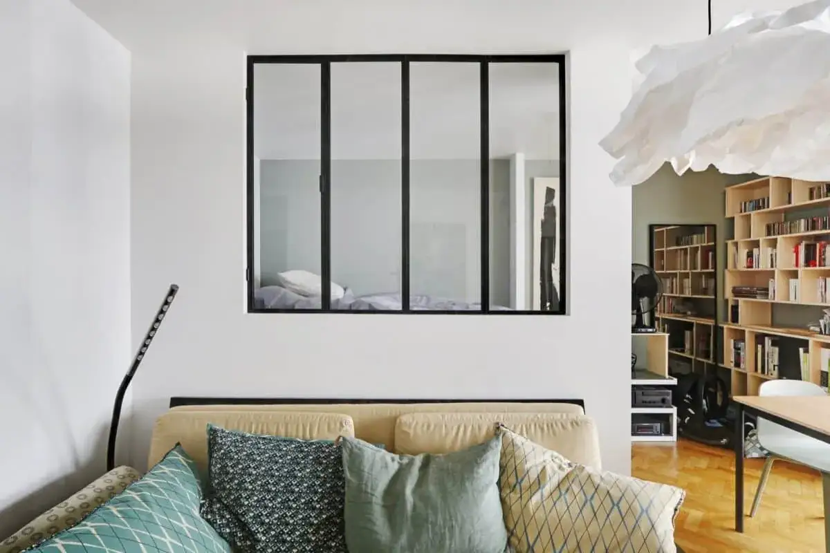 Fenêtre industrielle noire, vue sur une chambre avec lit, installée dans un mur blanc au-dessus d'un canapé.