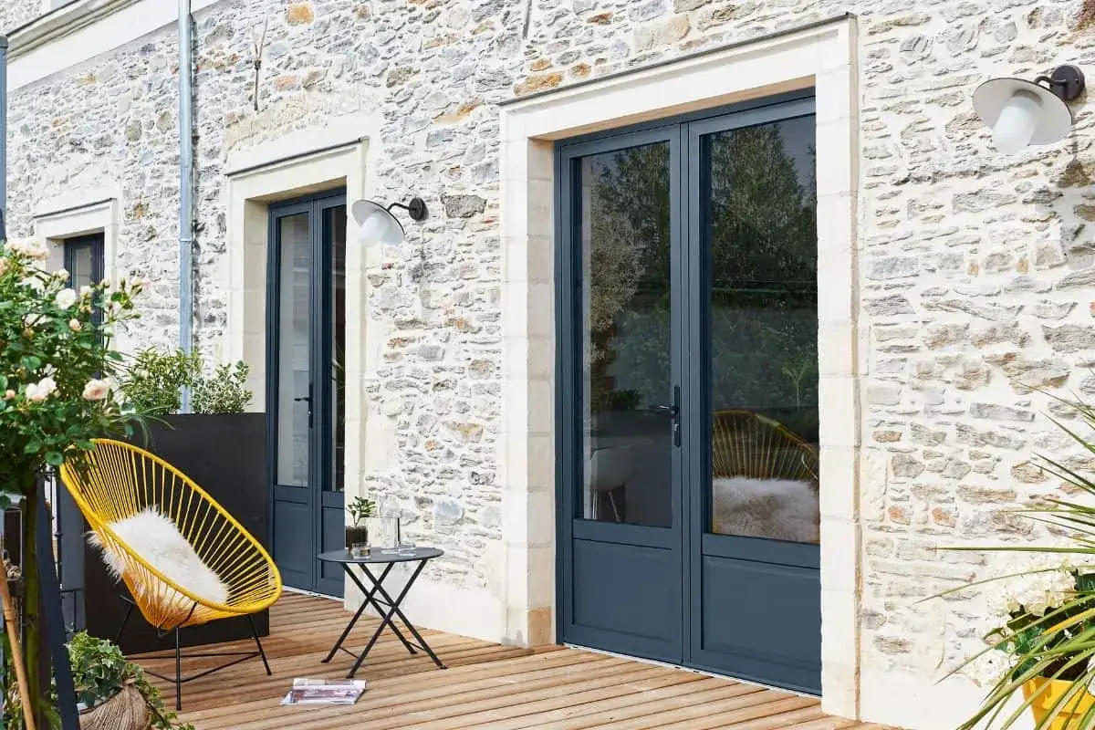 Une porte fenêtre qui s'ouvre vers l'extérieur, sur une terrasse en bois, avec une chaise jaune et une table.
