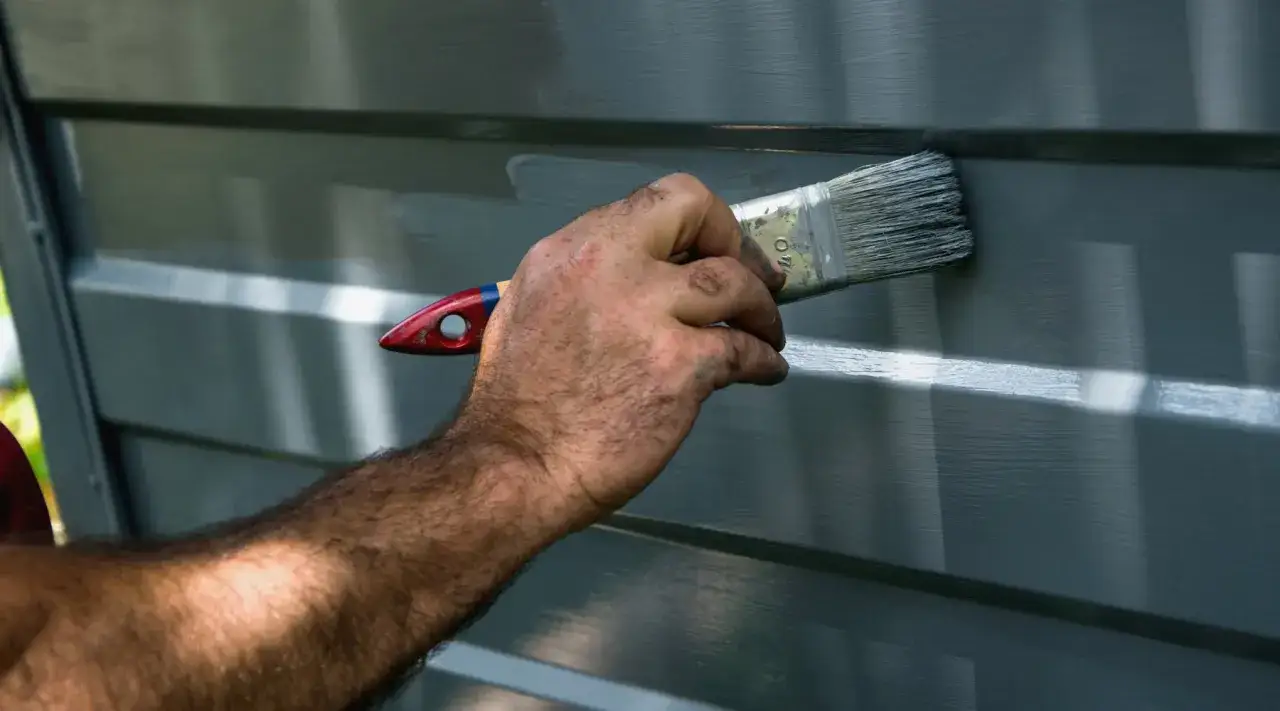 Main d'un peintre appliquant une couche de peinture grise sur une porte de garage.