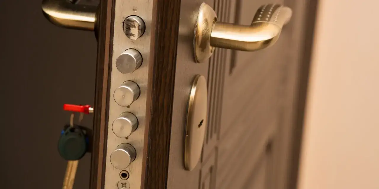 Poign&eacute;e dor&eacute;e et serrure de s&eacute;curit&eacute; sur une porte en bois sombre, avec des cl&eacute;s rouges et vertes. Un signe d'effraction porte est visible sur le cylindre de serrure.