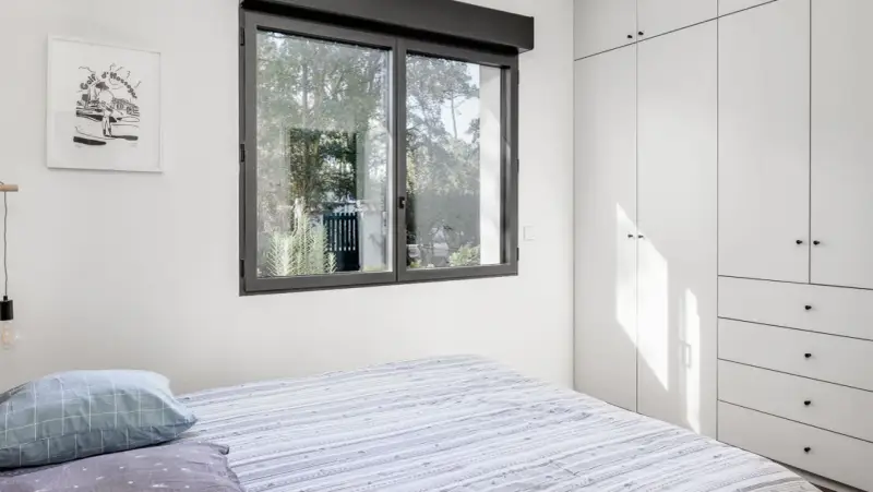 Une **ouverture fenêtre** laisse entrer la lumière naturelle dans une chambre moderne avec un lit à rayures et une armoire blanche.